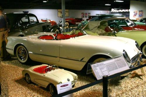 A vintage white convertible car with red interior displayed in a museum, alongside a small model car.