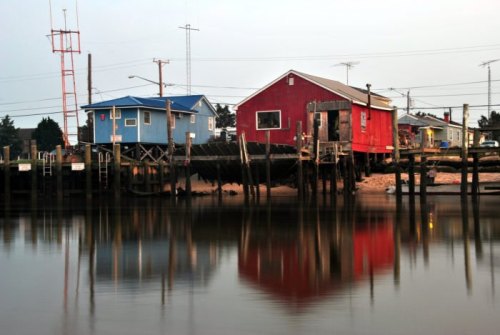 Colorful houses on stilts reflect in calm water at dusk, with power lines and a tower in the background.