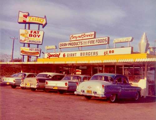 A vintage drive-in restaurant with classic cars parked outside, featuring a sign advertising giant burgers for $1.