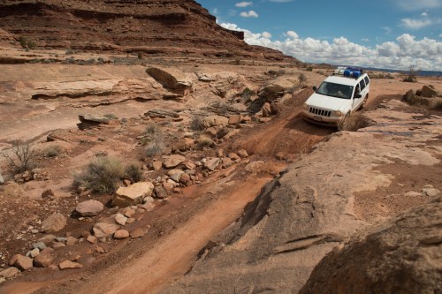A white SUV navigates a rocky, uneven terrain in a desert landscape under a blue sky with scattered clouds.