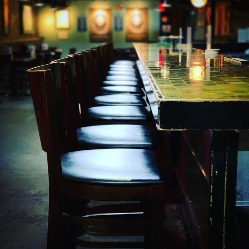 A row of empty wooden bar stools lined up at a dimly lit bar with a green tiled countertop.