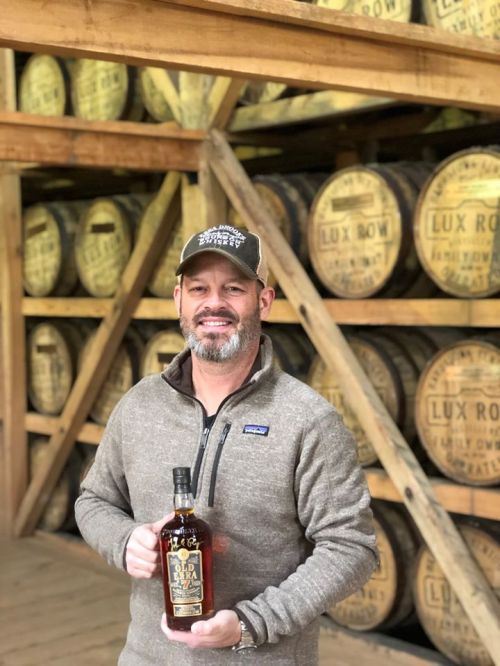 A man in a sweater and cap holds a bottle of whiskey in a distillery with wooden barrels in the background.
