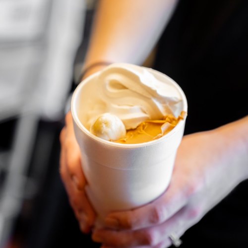 A person holds a cup filled with creamy ice cream topped with whipped cream and a dollop of caramel sauce.