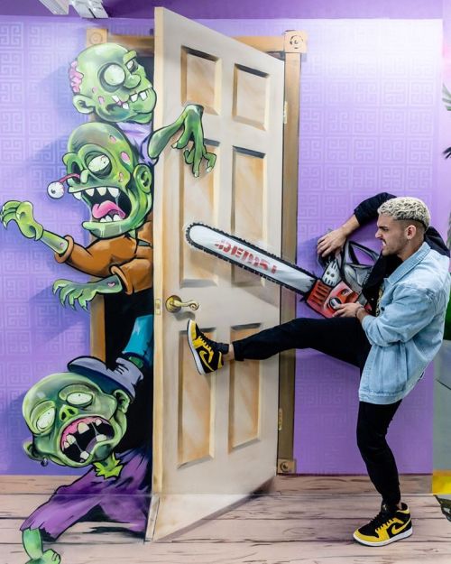 A person kicks open a door with a chainsaw, revealing cartoon zombies painted on the wall behind it.