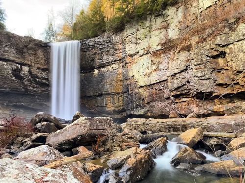 A serene waterfall cascades down rocky cliffs, surrounded by lush trees and smooth river stones.