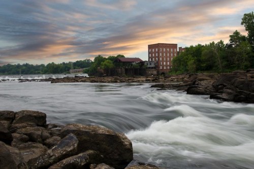 A serene river scene with flowing water, rocky shore, and a historic building under a colorful sky at sunset.