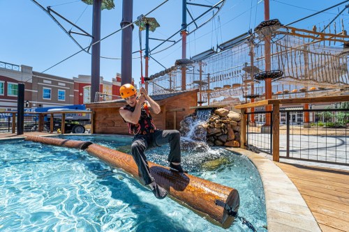 A person in a helmet balances on a log over water at an adventure park, with ropes and a climbing structure in the background.