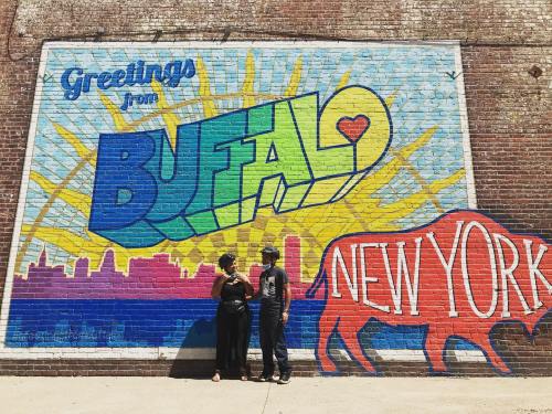 Two people stand in front of a colorful mural that says "Greetings from Buffalo, New York," featuring a red buffalo.