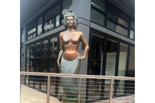 A tall, bronze mermaid statue stands outside a modern building, showcasing a stylized design and elegant posture.