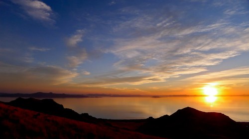A serene sunset over a calm lake, with silhouettes of hills and colorful clouds in the sky.