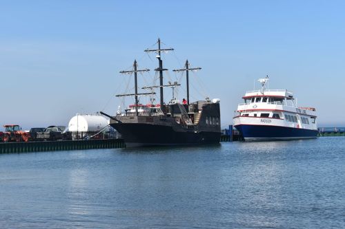 A black pirate ship and a white passenger ferry docked at a harbor under a clear blue sky.