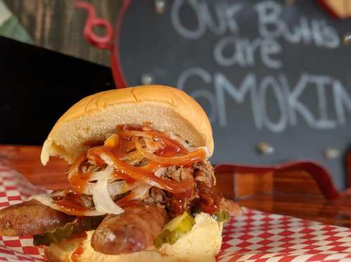 A close-up of a loaded sausage sandwich with onions, pickles, and barbecue sauce, with a sign in the background.