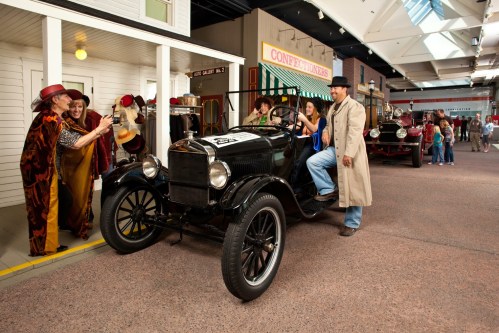 A vintage car in a museum setting, with people dressed in period costumes interacting and enjoying the scene.