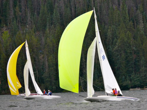 Two sailboats with colorful spinnakers race on a lake, surrounded by lush green trees in the background.