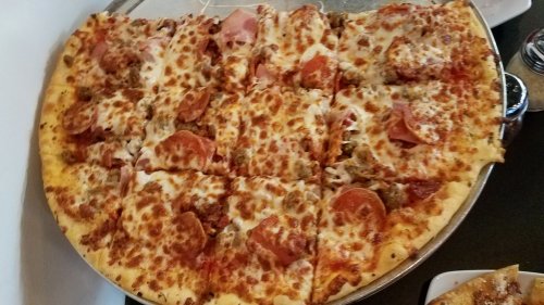 A large pizza topped with pepperoni, sausage, ham, and melted cheese, cut into square slices on a metal tray.