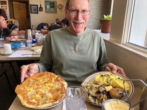 A smiling man holds two plates of food, one with a large omelet and the other with potatoes and a muffin, in a cozy diner.