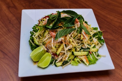 A fresh salad with shredded cabbage, vegetables, herbs, and lime wedges on a square plate.