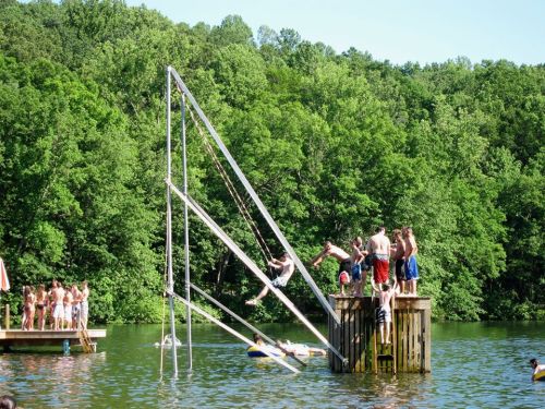 A group of children on a wooden platform by a lake, with a tall slide and others jumping into the water. Lush greenery surrounds.