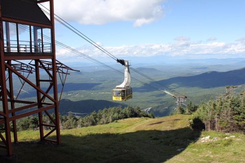 A cable car travels along a cable above a lush green landscape, with mountains and a blue sky in the background.