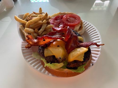 A juicy burger with two cheese slices, bacon, lettuce, and tomato, served with a side of crispy fries on a paper plate.