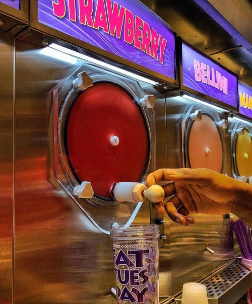 A hand holds a cup under a slushy machine dispensing strawberry slush, with colorful flavor labels above.
