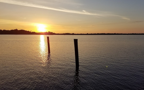 Sunset over calm water with two wooden posts silhouetted in the foreground. Soft colors reflect on the surface.