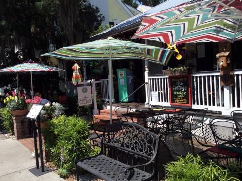 A charming café entrance with colorful umbrellas, outdoor seating, and vibrant flowers in a sunny setting.