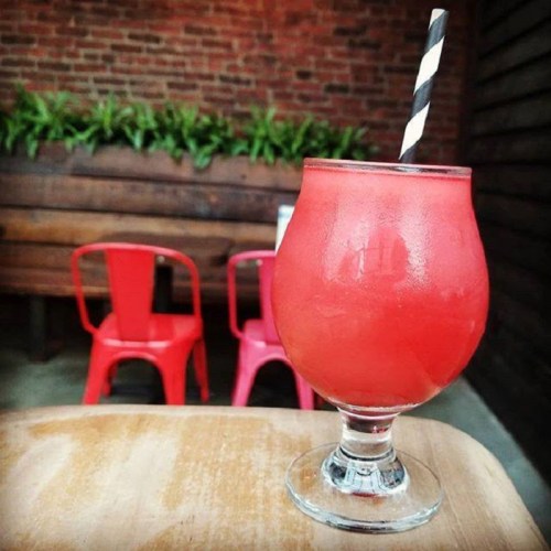 A vibrant red cocktail in a glass with a black and white striped straw, set on a wooden table with chairs in the background.