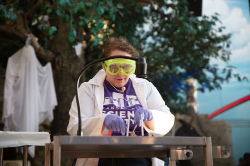 A person in a lab coat and goggles conducts an experiment at a science event, focused on their work.