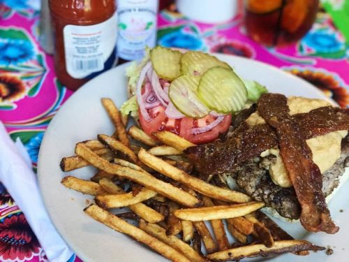 A plate of a burger topped with bacon, lettuce, tomato, pickles, and fries, set on a colorful floral tablecloth.