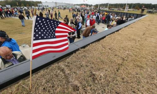 A crowd gathers near a memorial wall, with a small American flag in the foreground on a grassy area.