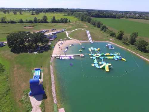 Aerial view of a water park with inflatable attractions on a lake, surrounded by green fields and trees.