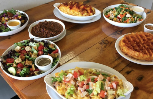 A wooden table with various dishes: salads, mac and cheese, chili, and waffles, all served in white bowls.