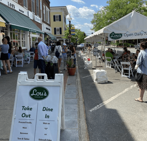 A busy street scene with outdoor dining at Lou's, featuring a sign for takeout and tables under a tent.
