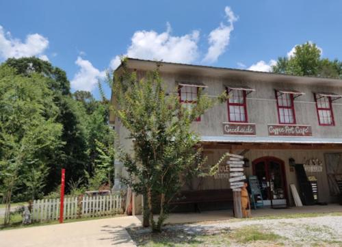 A rustic café building with red windows and signs, surrounded by greenery and a gravel path. Bright blue sky above.