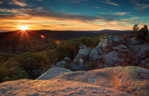 A stunning sunset over a rocky landscape, with a person standing on a cliff surrounded by lush trees and hills.