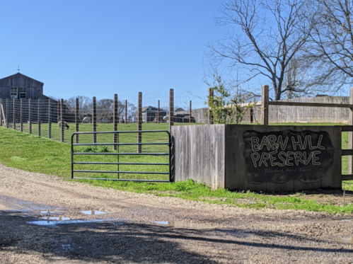 A rustic entrance to Barn Hill Preserve, featuring a wooden gate and a grassy area with a barn in the background.