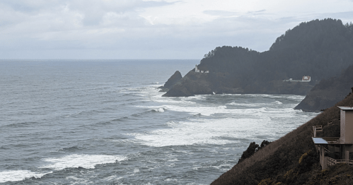 A coastal view with rolling waves, rocky cliffs, and a distant lighthouse under a cloudy sky.