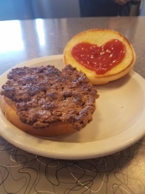 A hamburger bun with a heart-shaped ketchup design on the top half, and a cooked beef patty on a plate.