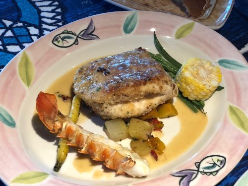 A plated dish featuring fish with sauce, lobster tail, corn, and vegetables on a decorative plate.