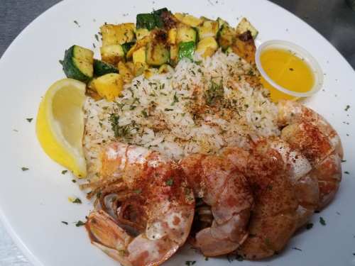 A plate of shrimp with rice, grilled zucchini, and a lemon wedge, served with a small container of sauce.