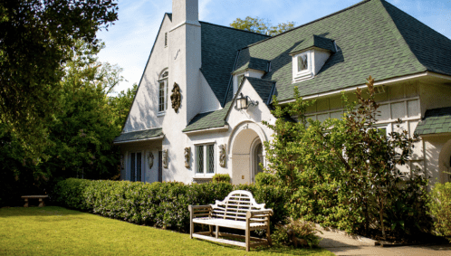 A charming white house with a green roof, surrounded by lush greenery and a wooden bench in the front yard.
