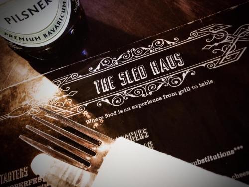 Close-up of a menu titled "The Sled Haus" with a fork and a bottle of beer in the background.