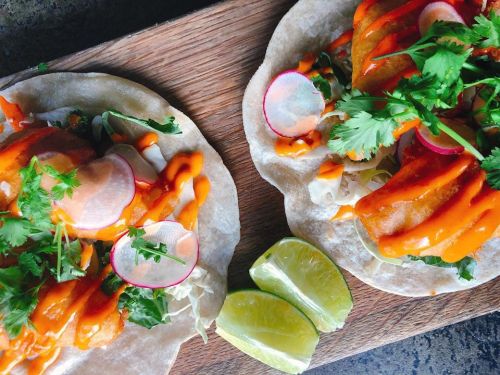 Two tacos on a wooden board, topped with cilantro, radishes, and drizzled with orange sauce, alongside lime wedges.