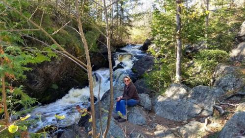 A person sits on a rock by a rushing river, surrounded by trees and rocky terrain in a natural setting.