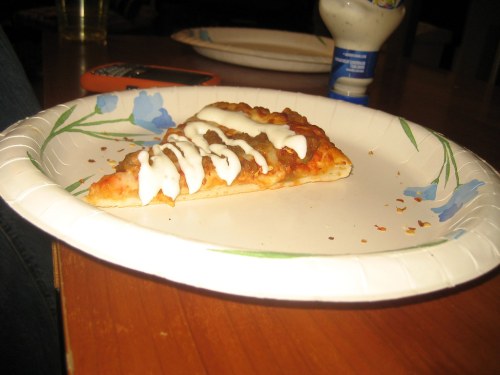 A slice of pizza on a paper plate, drizzled with white sauce, resting on a wooden table.