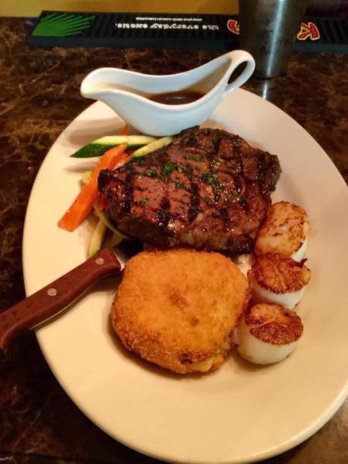 A plate featuring a grilled steak, scallops, a breaded croquette, and mixed vegetables, with a sauce on the side.