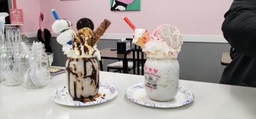 Two colorful milkshakes in jars topped with whipped cream, candies, and cookies, served on paper plates.