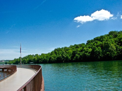 A serene river scene with a curved walkway, lush green trees, and a clear blue sky with a few clouds.