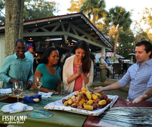 A group of four friends joyfully gathers around a table with a seafood feast, surrounded by a lively outdoor setting.
